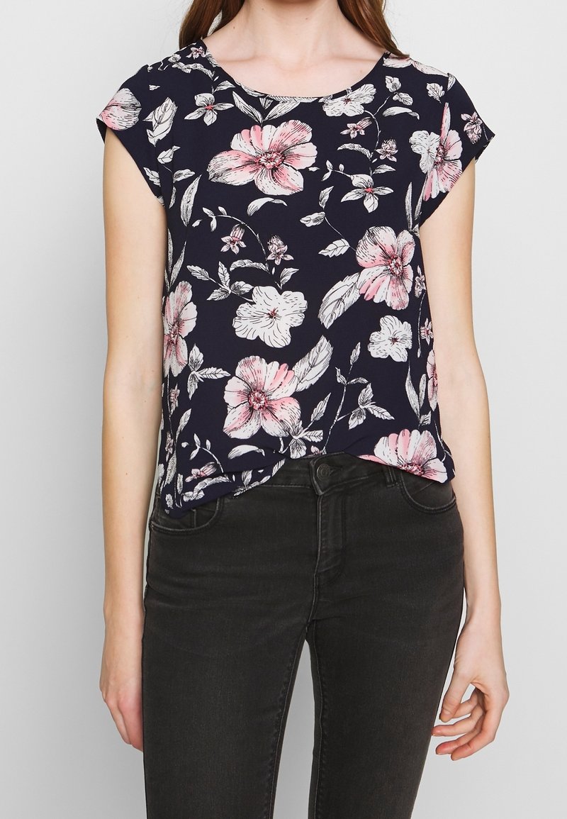 Femme portant un haut bleu marine à manches courtes avec des imprimés floraux roses et blancs, associé à un jean noir sur un fond uni.