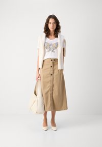 Hvit t-skjorte med en fargerik vingsdesign, kombinert med en beige midi-skjørt med frontknapper, kremfarget cardigan og lyse hæler.
