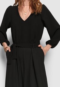 Femme portant une robe noire à col en V avec des manches longues, une ceinture à la taille nouée, les mains dans les poches, sur un fond uni.