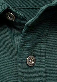 Col de chemise en denim vert avec un bouton-pression argenté, tissu texturé et motif à côtes horizontales, présentant un détail de couture.