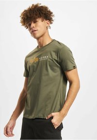 Bombažna majica olivno zelene barve z okroglim ovratnikom, kratkimi rokavi in rumenim grafičnim logotipom na prsih. Gladek tekstura, standardni kroj.