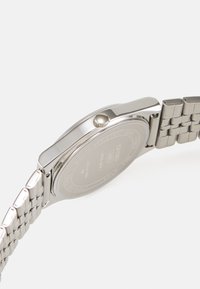 Montre en acier inoxydable argenté avec un boîtier rond, une couronne texturée et un bracelet en métal comportant des maillons plats, vue de côté.