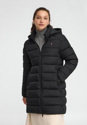 Donna che indossa un cappotto imbottito trapuntato nero con cappuccio sopra un dolcevita grigio e gonna crema, in piedi con una mano in tasca davanti a uno sfondo bianco.