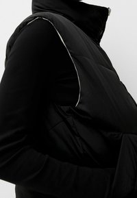 Black puffer vest with a high collar, featuring a smooth texture and white piping along the armholes. Front zipper closure is visible.