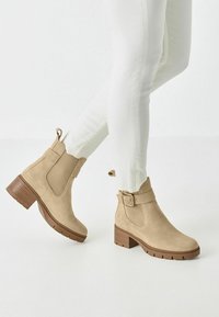 Bottines en daim beige avec un talon épais, accent en boucle et panneaux latéraux élastiques, associées à un jean slim blanc. Semelle en caoutchouc plate.