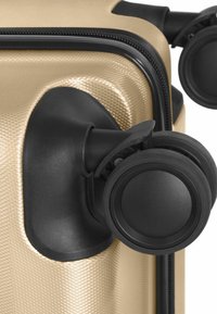 Gros plan sur deux roues pivotantes noires fixées au coin inférieur d'une valise rigide texturée couleur or.
