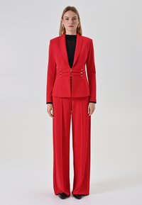 Rode op maat gemaakte pak met een aansluitende blazer met een diepe revers en dubbele gespsluitingen, gecombineerd met wijde broekspijpen. Gladde stoftextuur.