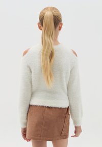 Weißer, flauschiger Pullover mit offenen Schultern, kombiniert mit einem braunen Cordminirock mit Seitentasche. Glatte Textur und gemütliches Design.