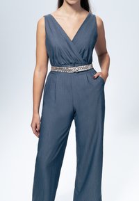 Denim jumpsuit med dyb V-udskæring og brede ben, udstyret med et sølvfarvet fletbælte og sidelommer. Blød tekstur og flot blå farve.