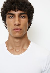 Weißes Rundhals-T-Shirt aus glattem Stoff, körpernah geschnitten, getragen von einer Person mit lockigem schwarzen Haar und neutralem Ausdruck.