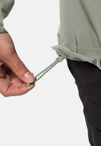 Pullover verde chiaro, strutturato, con coulisse regolabile all'orlo; mano che stringe un bottone per stringere; pantaloni scuri visibili.