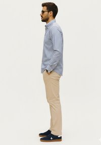 Camisa de botón azul a rayas, pantalones beige ajustados, zapatillas navy y gafas de sol. La tela parece suave con un corte entallado.