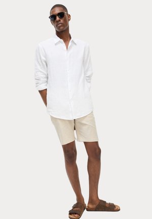 White linen long-sleeve shirt, beige shorts, brown sandals. Slim fit, button closure, relaxed design, model stands with hands in pockets.