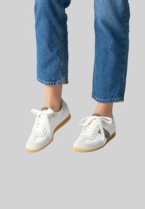 Person wearing cropped blue jeans and white casual sneakers with gum soles against a gray background.