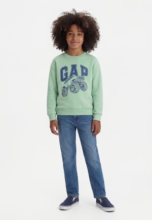 Bambino con capelli ricci che indossa una felpa verde chiaro con un monster truck e la scritta "GAP", jeans blu e scarpe slip-on blu navy, in piedi.