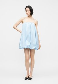 Jeune femme debout portant une mini-robe bleu clair à ourlet bouffant avec de fines bretelles et des ballerines noires pointues, sur un fond blanc uni.