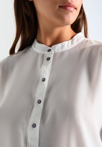Blusa bianca con colletto alla mandarino, tessuto testurizzato e una patta con sei bottoni rotondi scuri per la chiusura.