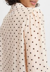 Blusa pescoce pallido con pois neri, tessuto testurizzato, maniche a sbuffo e colletto classico, con delicate pieghe e movimento.