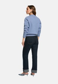 Donna in un maglione azzurro chiaro, jeans scuri con risvolti e décolleté blu kitten, si trova di spalle, guardando verso sinistra su uno sfondo semplice.