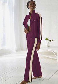 Tummanpunainen collegepuku, jossa on vaaleanväriset sivuraitoja; vetoketjullinen takki ja siihen sopivat housut. Pehmeä kangas, rento istuvuus, yhdistetty valkoisen paidan kanssa.