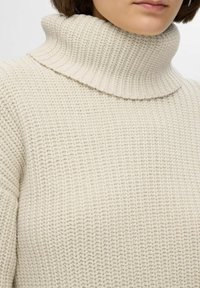 Teksturirana krema turtleneck pulover s visokim rebrastim ovratnikom in ohlapnim pletenjem. Mehka, udobna tkanina z sproščenim krojem.