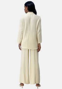 Lys cremefarvet to-delt sæt med tekstur, bestående af en oversized jakke med lange ærmer og bukser med vide ben, kombineret med hæle.