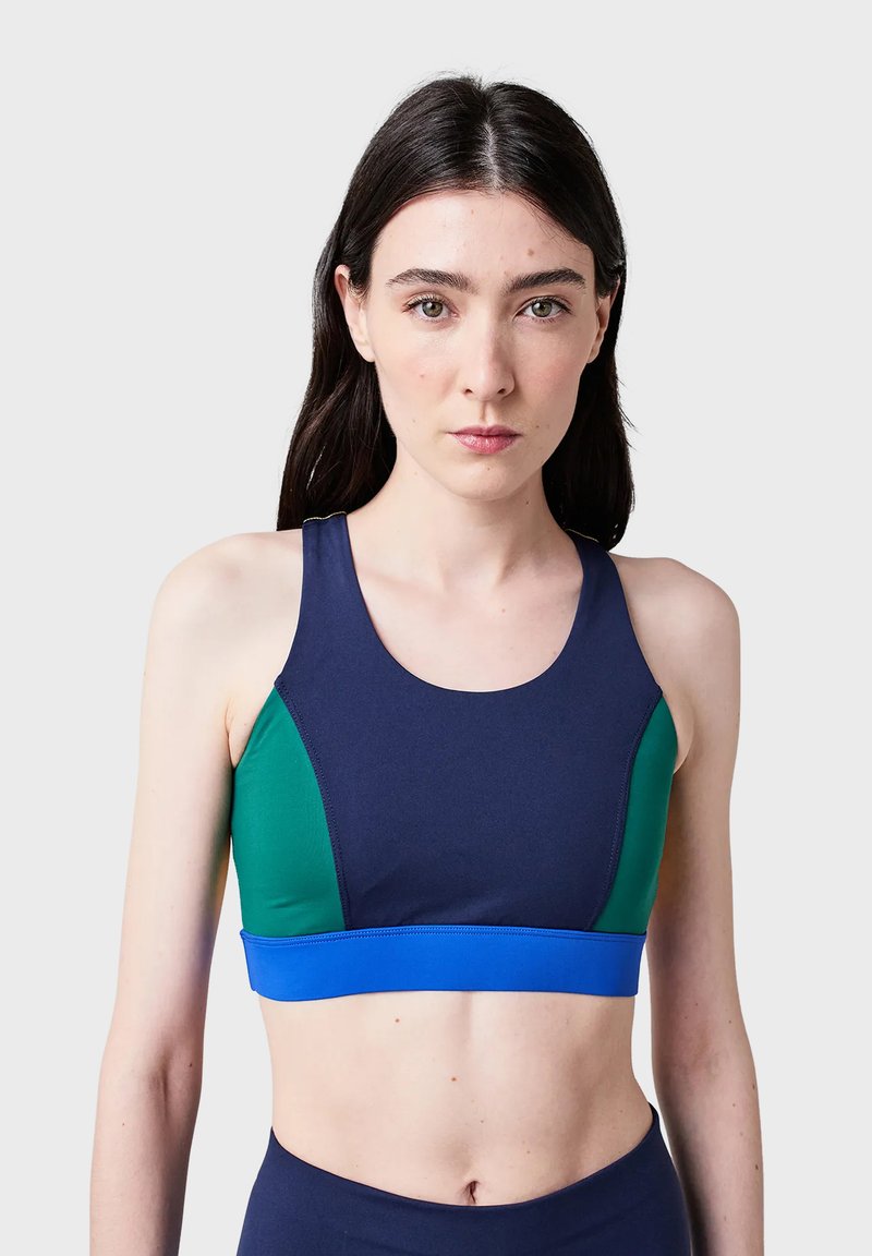 Ung kvinna med långt mörkt hår klädd i en marinblå, grön och blå färgblockerad sport-BH, vänd framåt mot en enfärgad bakgrund.