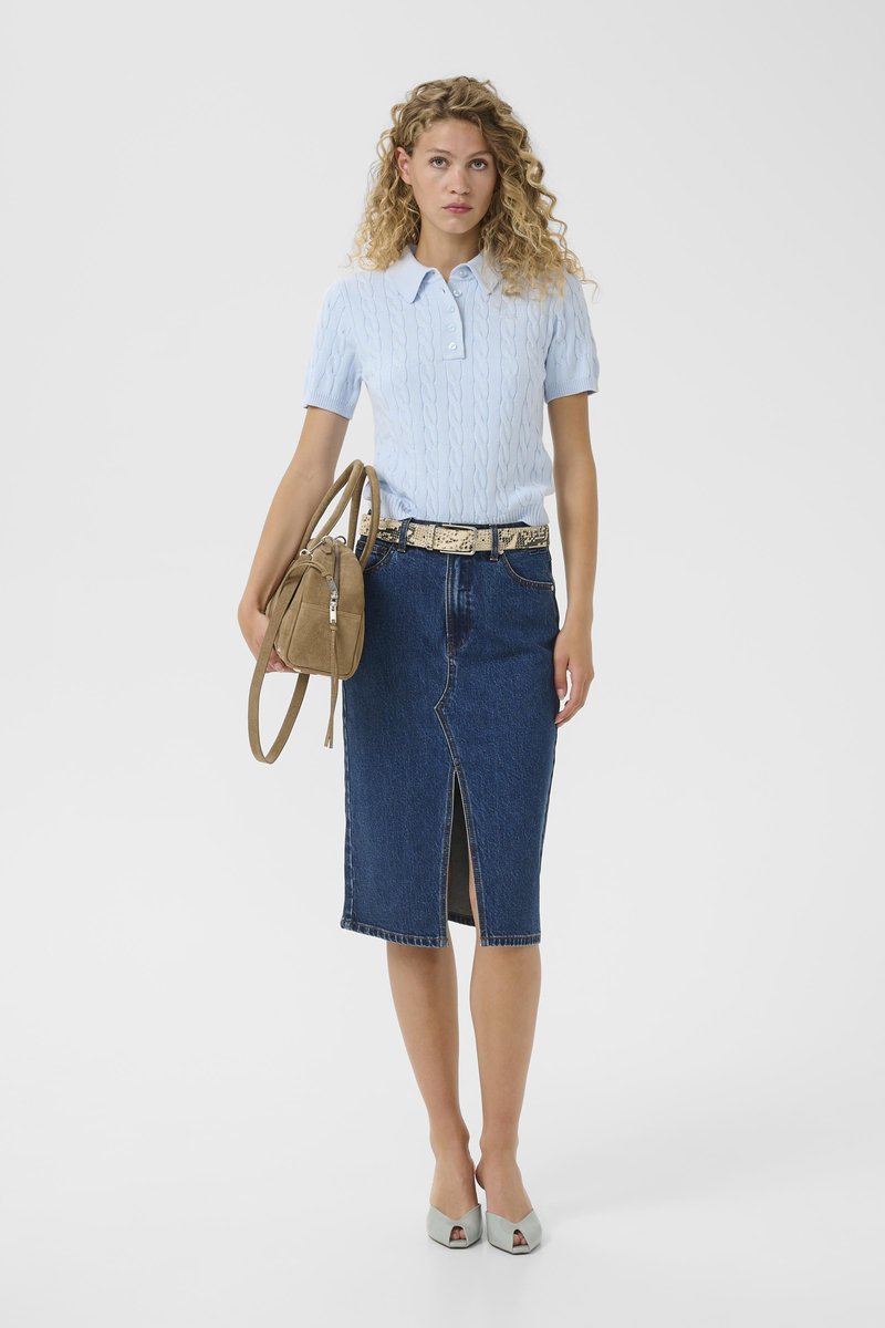 Femme portant un pull léger à manches courtes bleu clair, une jupe en denim foncé longueur genou avec une fente à l'avant, une ceinture beige, des talons blancs ouverts, tenant un sac à main marron clair.