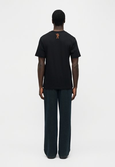 Άνδρας που στέκεται με την πλάτη γυρισμένη, φορώντας απλό μαύρο κοντομάνικο μπλουζάκι με μικρό πορτοκαλί γραφικό αστροναύτη στην άνω πλάτη και σκούρο παντελόνι με φαρδιά μπατζάκια.