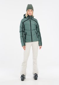 Chaqueta de plumas verde con acentos negros y bolsillos con cremallera, combinada con pantalones de color claro y botas de esquí negras. El modelo lleva un gorro de lana.