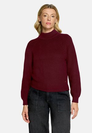 Femme blonde portant un pull à col roulé côtelé rouge foncé et un jean noir taille haute, debout devant un fond clair.