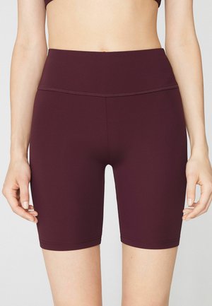 Mujer con pantalones cortos de ciclista ajustados de cintura alta y color púrpura oscuro, mostrada desde la mitad del torso hasta la mitad del muslo sobre un fondo liso.