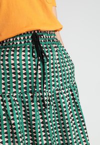 Jupe à motifs verts et noirs sur fond rose, avec des rayures verticales et des formes triangulaires, taille élastique et cordon de serrage.