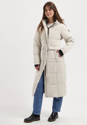 Frau trägt einen langen, hellbeigen, gesteppte Mantel mit Kapuze, blaue Jeans und schwarze Stiefeletten, steht mit einer Hand in der Tasche.