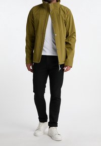 Man i en olivgrön jacka över en vit skjorta, svarta byxor och vita sneakers som står mot en enkel vit bakgrund.