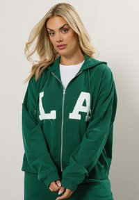 Felpa verde con zip realizzata in tessuto morbido, caratterizzata da grandi lettere bianche "LA", cappuccio con cordino e polsini a costine. Accentuata da tasche laterali.