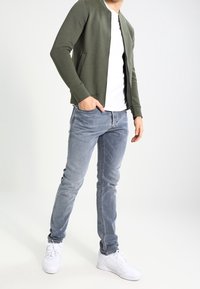 Chaqueta bomber acolchada verde oliva con una camiseta blanca, combinada con jeans azul claro y zapatillas blancas. Diseño simple, silueta ajustada.