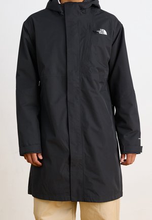 Schwarze, lange wasserdichte Kapuzenjacke mit Frontreißverschluss, Klappenverschluss und weißem The North Face-Logo auf der Brust, getragen mit beigen Hosen.