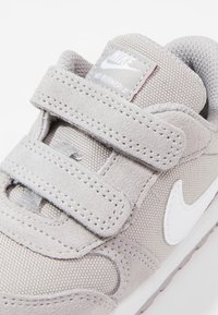 Světlé �šedé tenisky vyrobené z textilu a semiše, se suchými zipy, síťovanými detaily a bílým logem Nike swoosh. Viditelný texturovaný povrch.