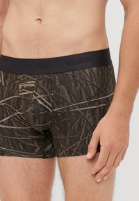Brajne kamuflažne bokserice s črnim elastičnim pasom, na katerem je vbočeno napisano "HOLLISTER". Oblikovanje vključuje teksture, ki posnemajo listje.