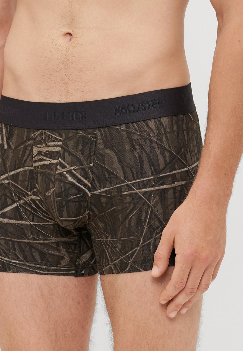 Brązowe bokserki w kamuflażu z czarnym elastycznym pasem, na którym jest wytłoczone "HOLLISTER". Projekt zawiera tekstury przypominające liście.
