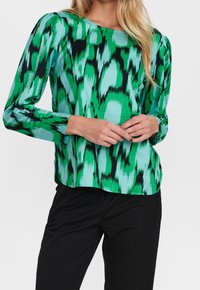 Blouse à manches longues verte et turquoise avec un motif abstrait audacieux. Tissu doux et coupe décontractée, associée à un pantalon sombre.
