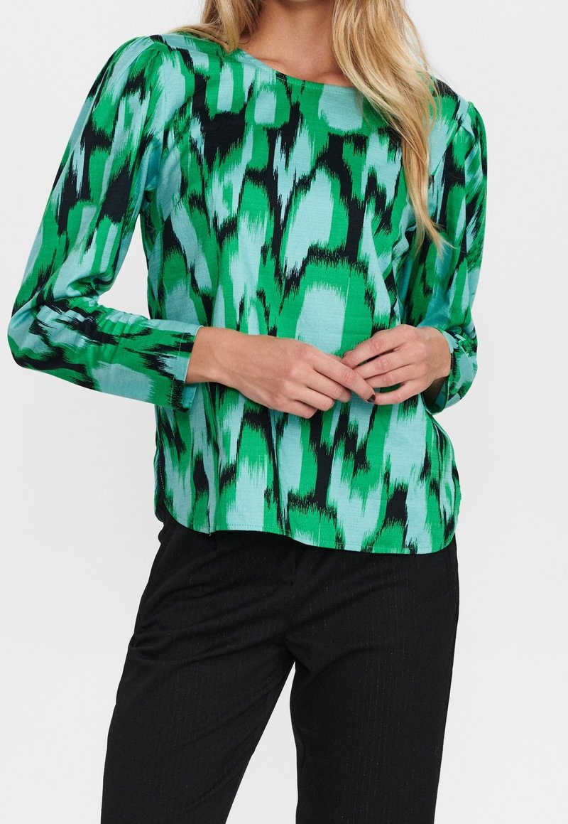 Blouse à manches longues verte et turquoise avec un motif abstrait audacieux. Tissu doux et coupe décontractée, associée à un pantalon sombre.