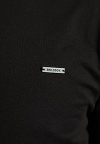 Czarna bawełniana koszulka z małą srebrną metalową metką z napisem "SMILODOX" wytłoczonym. Gładka tekstura i dopasowany krój.