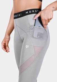 Pilki leggingai su juodu juosmuo, ant kurio yra "MOROTAI" logotipas. Su šoniniu kišeniu telefonui, tinklinėmis detalėmis ir stilingu dizainu.