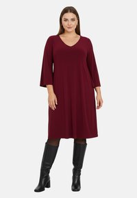 Robe bordeaux en tissu doux avec un décolleté en V, des manches à trois quarts évasées et une coupe en A. Associée à des bottes noires montantes.