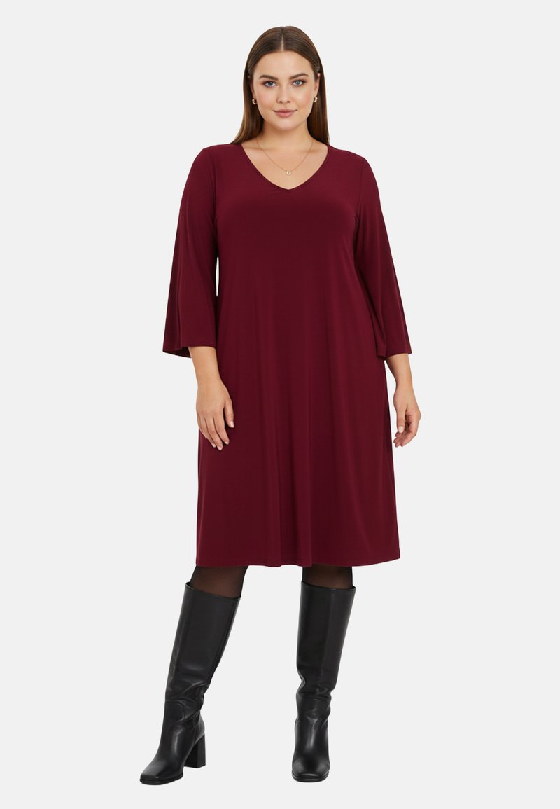 Robe bordeaux en tissu doux avec un décolleté en V, des manches à trois quarts évasées et une coupe en A. Associée à des bottes noires montantes.
