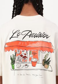 Cream t-shirt featuring a colorful illustration of a bistro with a red awning, plant pots, and café items like cups and pastries.