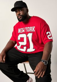 Rød t-shirt med korte ærmer, der har "NEW YORK" og "21" i hvid, parret med sorte bukser og en sort kasket. Sølvur på håndleddet.