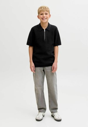 Nasmejan deček stoji in nosi črno polo majico s kratkimi rokavi, sive kavbojke in bele superge na enobarvnem svetlem ozadju.